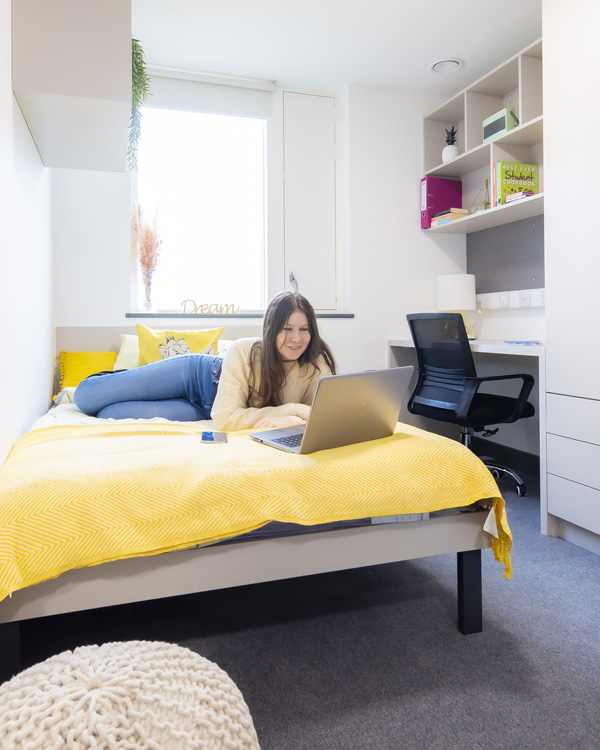 A female student is relaxing on the bed using her laptop in modern student accommodation