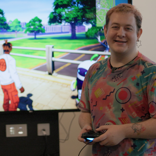 A student wearing a colourful tshirt is smiling and playing Fortnite on a console