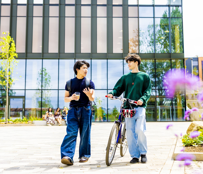 Two male students are walking around the Student Village. One has a coffee and the other male is pushing a bicycle