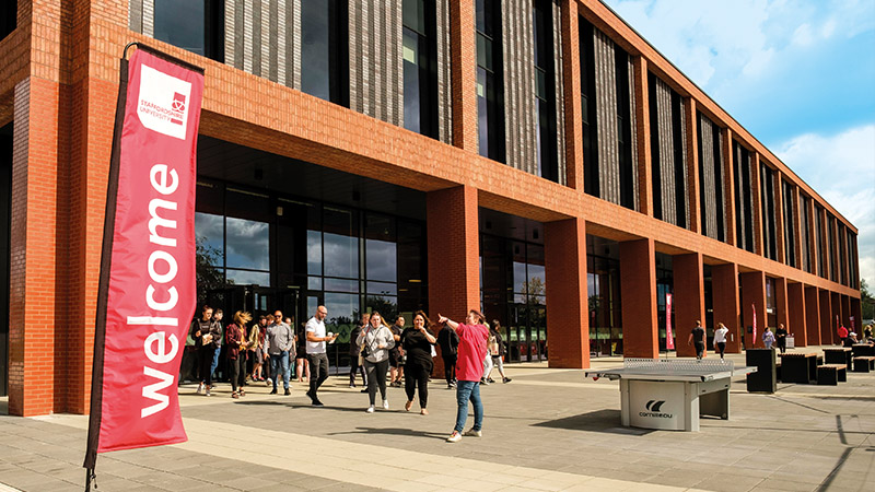 Student ambassador wearing a red Staffordshire University polo shirt directs a group of visitors during a campus tour outside our Catalyst building