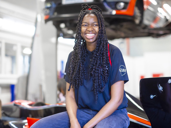 A female with long dark hair is smiling with a relaxed pose in the mechanics workshop