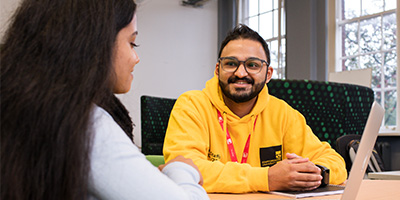 Male student chats to female student in our Thompson library. He is wearing a yellow hoody and student lanyard. She is wearing a grey sweater and black scarf.